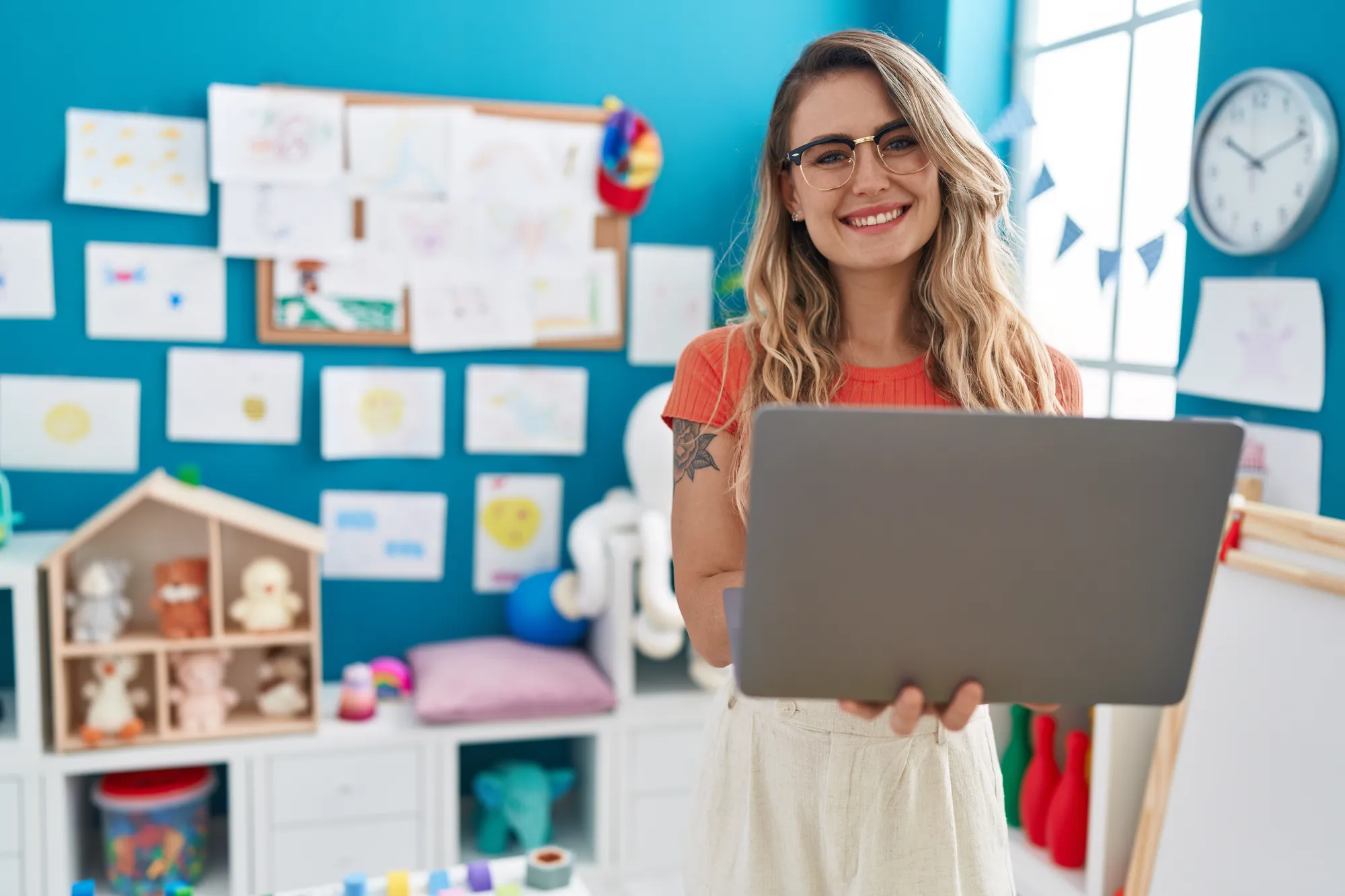 A smiling teacher holding a laptop in a bright classroom